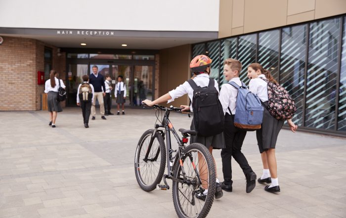 Group Of High School Students Wearing Uniform Arriving At School Walking Or Riding Bikes Being Greeted By Teacher